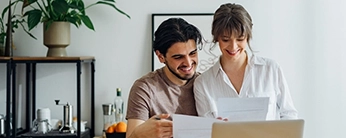 Jeune couple souriant, installé devant un ordinateurs, divers papiers en main